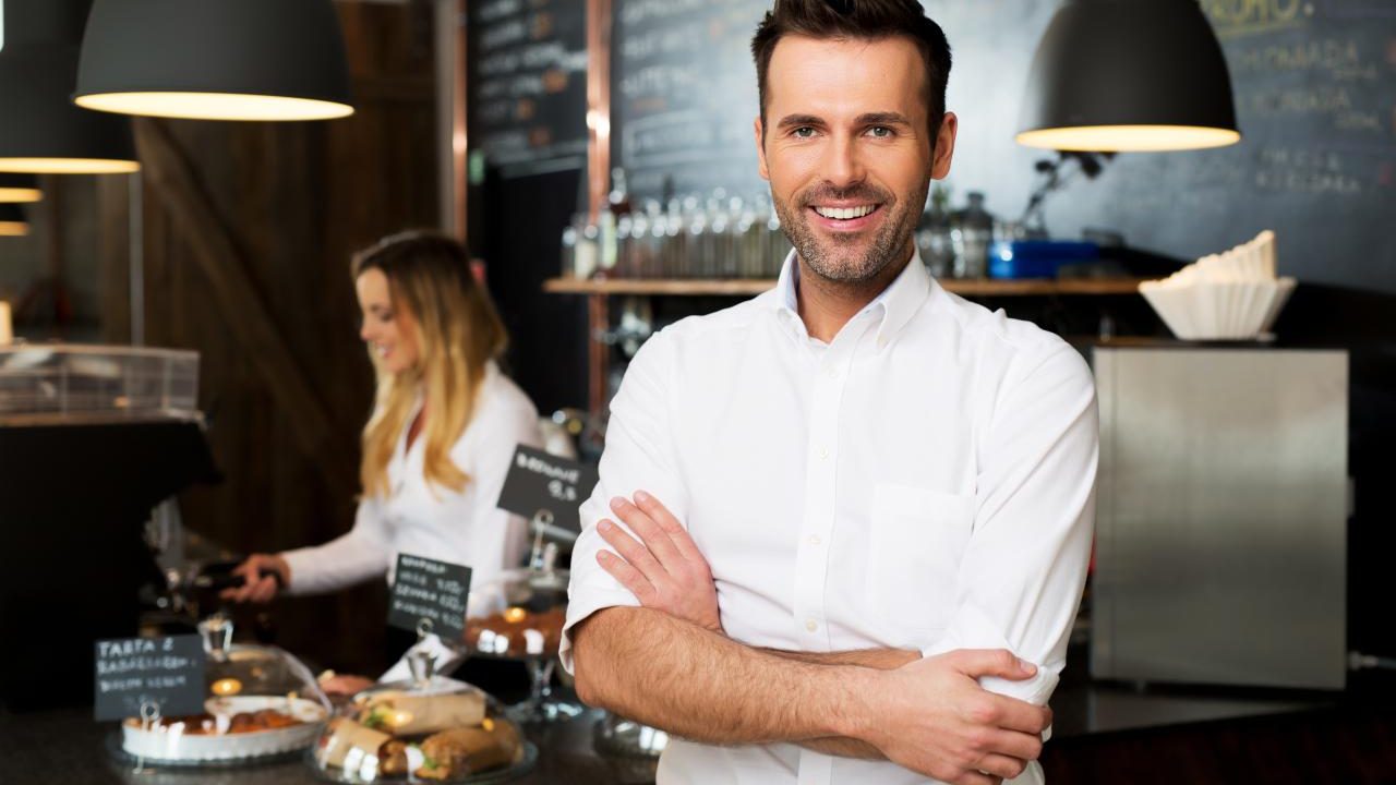 Happy small business owner standing at front of bar with employee in background preparing coffee